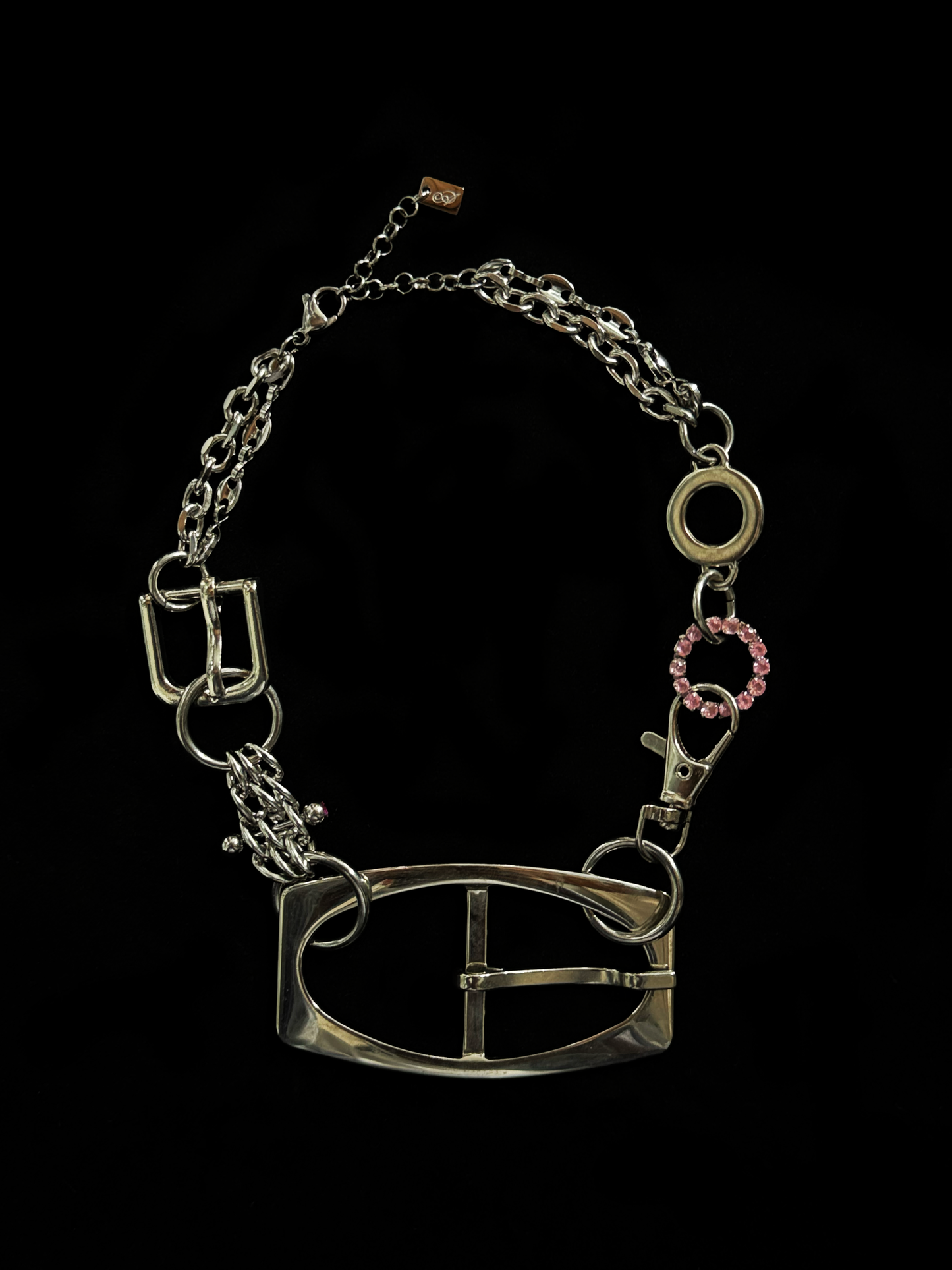A bold, industrial-inspired upcycled necklace featuring a repurposed equestrian-style buckles, layered chains, a piercing and a pop of pink rhinestones.