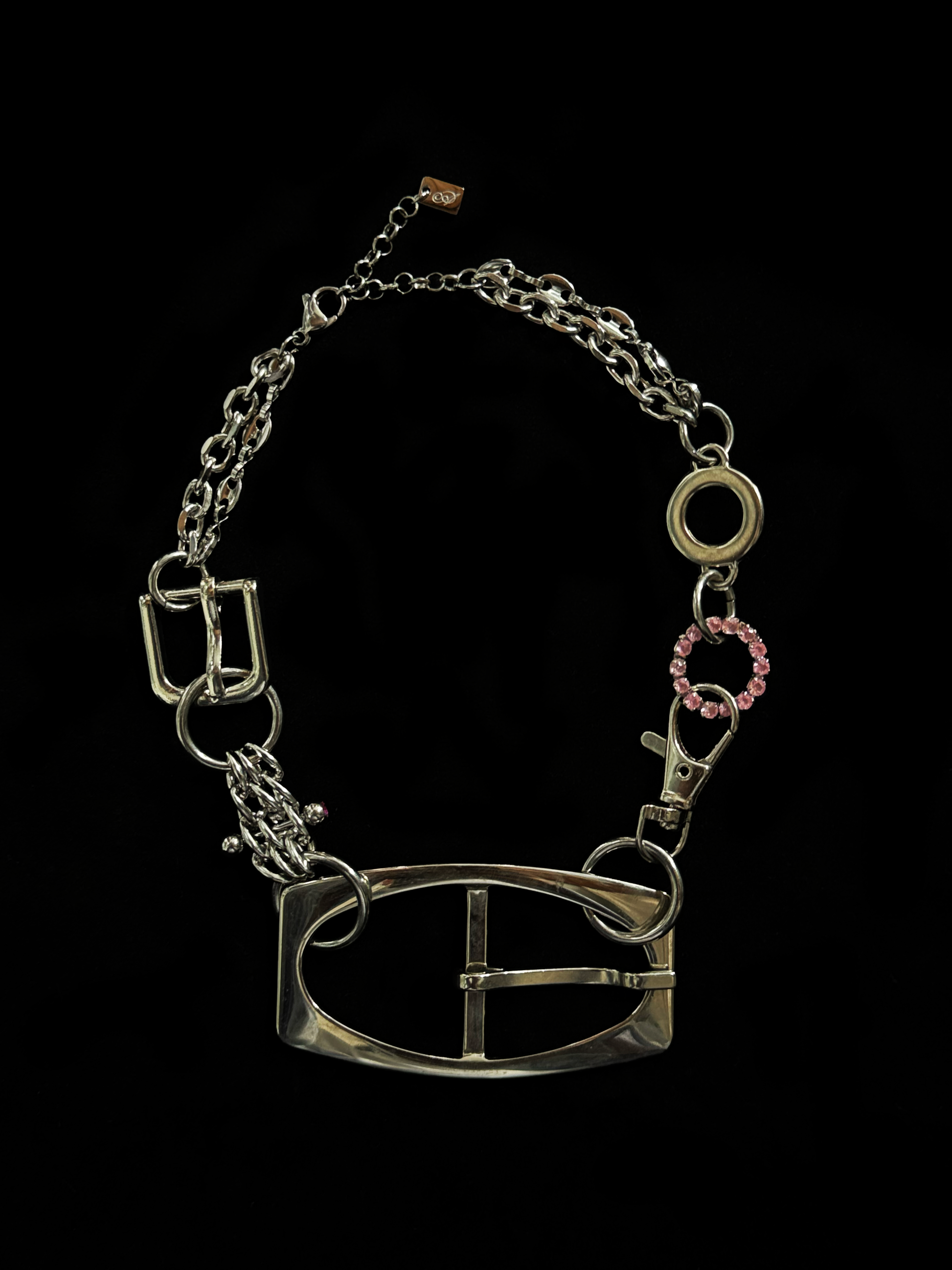 A bold, industrial-inspired upcycled necklace featuring a repurposed equestrian-style buckles, layered chains, a piercing and a pop of pink rhinestones.