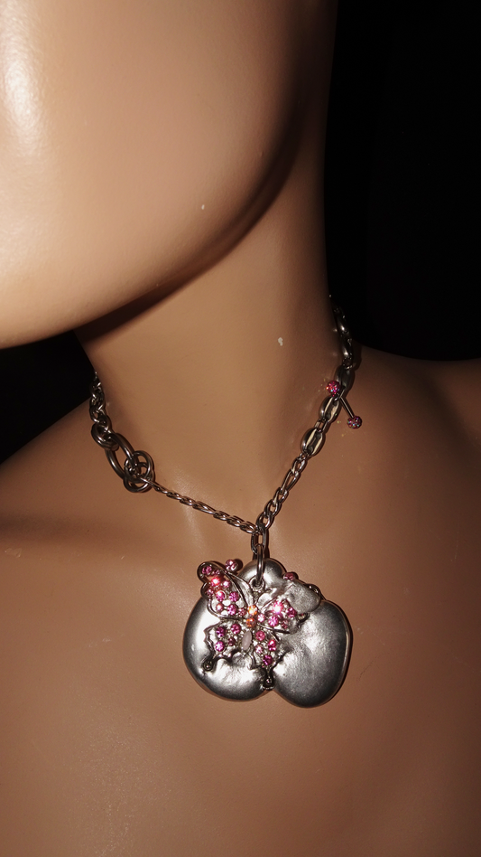 Upcycled asymmetrical necklace with pink rhinestone butterfly on organic silver-tone pendant, vintage chains, and unique piercing detail. Bold, sustainable statement jewelry.